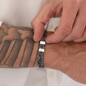 May include: A black and gray braided bracelet with two silver charms. The charms are engraved with the text "JOS" and "10.12.2022".