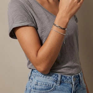 May include: A grey v-neck t-shirt and blue denim jeans paired with two bracelets. One bracelet is a thin black cord featuring small metallic beads, while the second is a delicate silver-toned chain with a textured, beaded appearance.