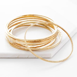 Può includere: Una collezione di braccialetti dorati con superficie strutturata. I braccialetti sono impilati su una superficie quadrata bianca. I gioielli sono presentati in un ambiente studio con illuminazione soffusa.