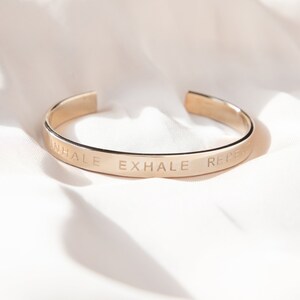 May include: A gold-colored cuff bracelet with the words "INHALE EXHALE REPEAT" engraved on the outside. The bracelet is open at the bottom and sits on a white, textured surface.