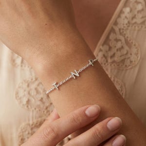 May include: A silver-toned chain bracelet featuring three individual capital letter charms spelling out the letters T, N, and P. The letters are evenly spaced along the delicate metal link chain, which wraps around the wrist with a polished finish.