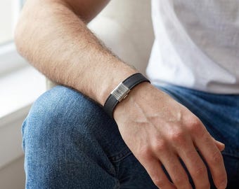 May include: Black leather bracelet with a silver clasp. The bracelet is worn on a person's wrist, showcasing its simple and elegant design. The clasp appears to be made of polished metal, adding a touch of sophistication to the accessory.