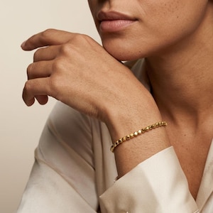 May include: A gold-coloured beaded bracelet is worn on a wrist. The bracelet is made of small, faceted beads. The person is wearing a cream-coloured satin shirt. The bracelet is a simple, elegant piece of jewellery.
