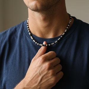 Pode incluir: Um colar de contas com contas prateadas e pretas alternadas. O colar é usado sobre uma camiseta azul escura. O fecho é prateado. Um acessório simples, mas elegante.
