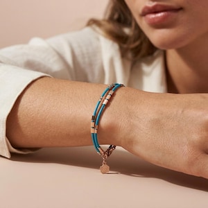 Può includere: Un braccialetto turchese e oro rosa con un design a doppio filo. Il braccialetto presenta piccoli accenti rettangolari in oro rosa e un ciondolo in oro rosa con un motivo floreale. Il braccialetto è indossato al polso.