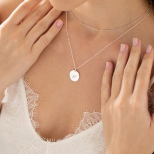 Peut inclure: Un collier en argent avec un pendentif rond gravé de la lettre "M". Le collier est superposé à une délicate chaîne perlée. Le bijou est porté par une personne vêtue d'un haut en dentelle blanche.