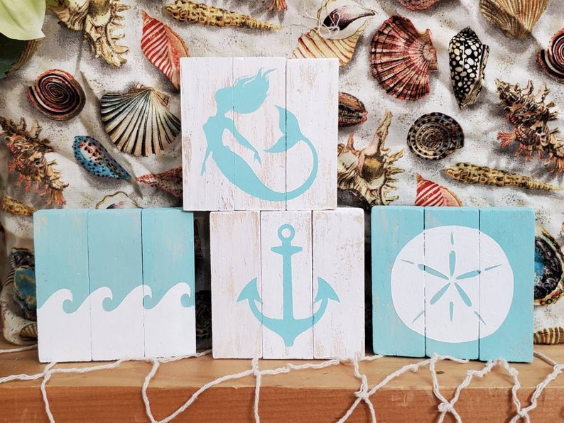K&ouml;nnte beinhalten: Drei Holzschildchen mit Strandthema. Das obere Schild zeigt eine blaue Meerjungfrau-Silhouette. Das mittlere Schild zeigt eine blaue Anker-Silhouette. Das untere Schild zeigt eine wei&szlig;e Sanddollar-Silhouette.