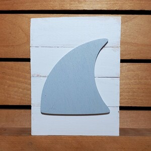 May include: A white wooden sign with a light blue shark fin cutout. The sign is made of reclaimed wood and has a rustic, farmhouse style.