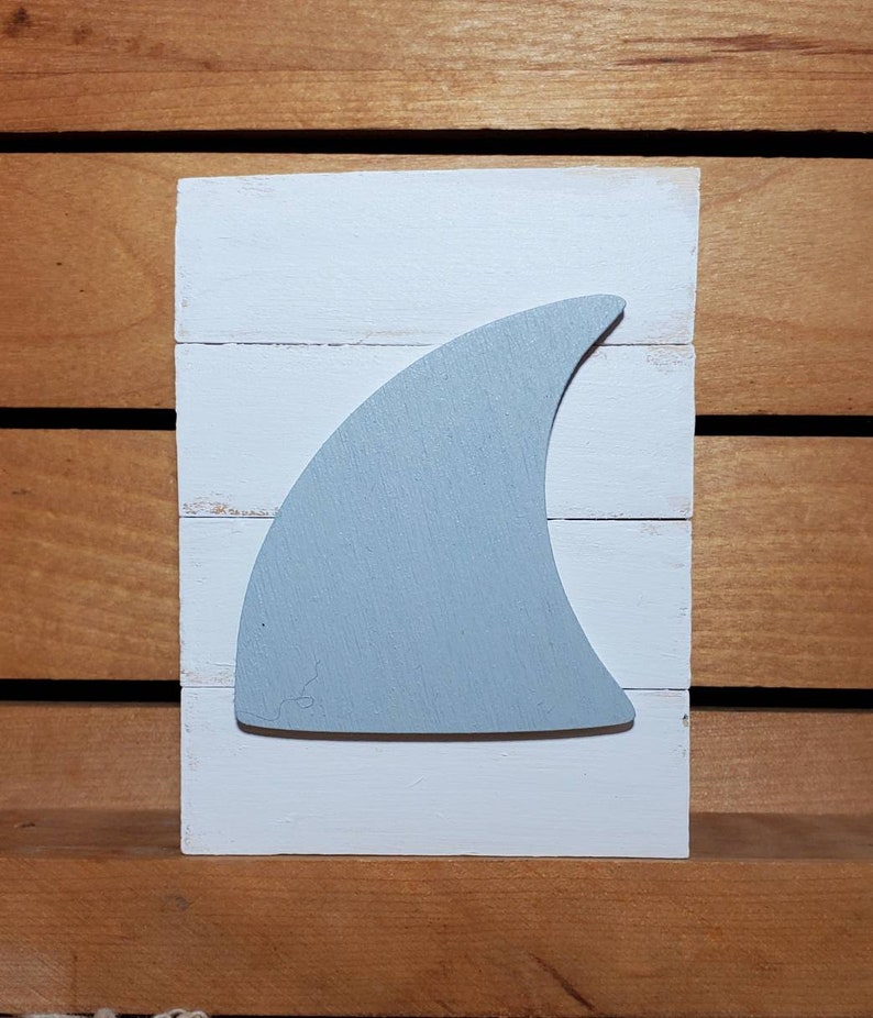 May include: A white wooden sign with a light blue shark fin cutout. The sign is made of reclaimed wood and has a rustic, farmhouse style.