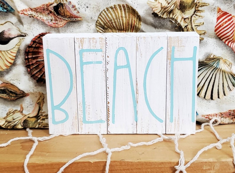 K&ouml;nnte beinhalten: Ein wei&szlig;es Holzschild mit dem Wort "BEACH" in hellblau bemalt. Das Schild besteht aus einzelnen Holzbrettern.