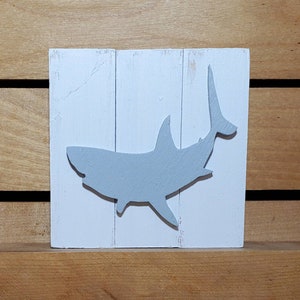 May include: A white wooden sign with a grey silhouette of a shark. The shark is facing to the right and has its tail raised in the air.