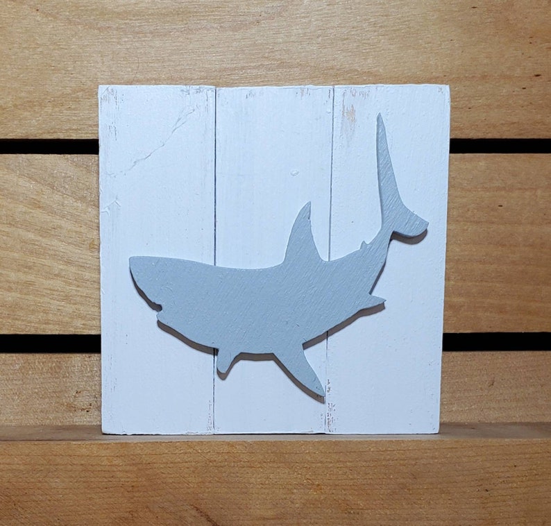 May include: A white wooden sign with a grey silhouette of a shark. The shark is facing to the right and has its tail raised in the air.