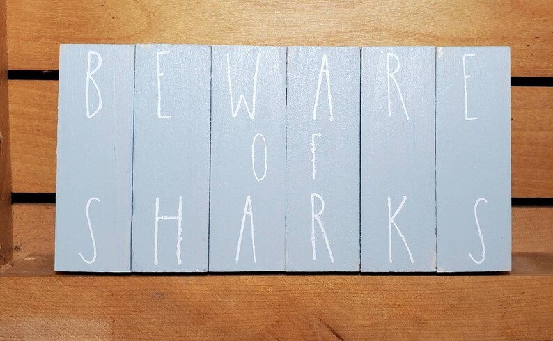 May include: A light blue wooden sign with the words "BEWARE OF SHARKS" written in white paint.