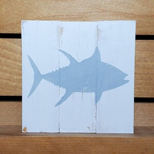 May include: A light blue silhouette of a tuna fish painted on a white wooden sign. The sign is made of three pieces of wood.