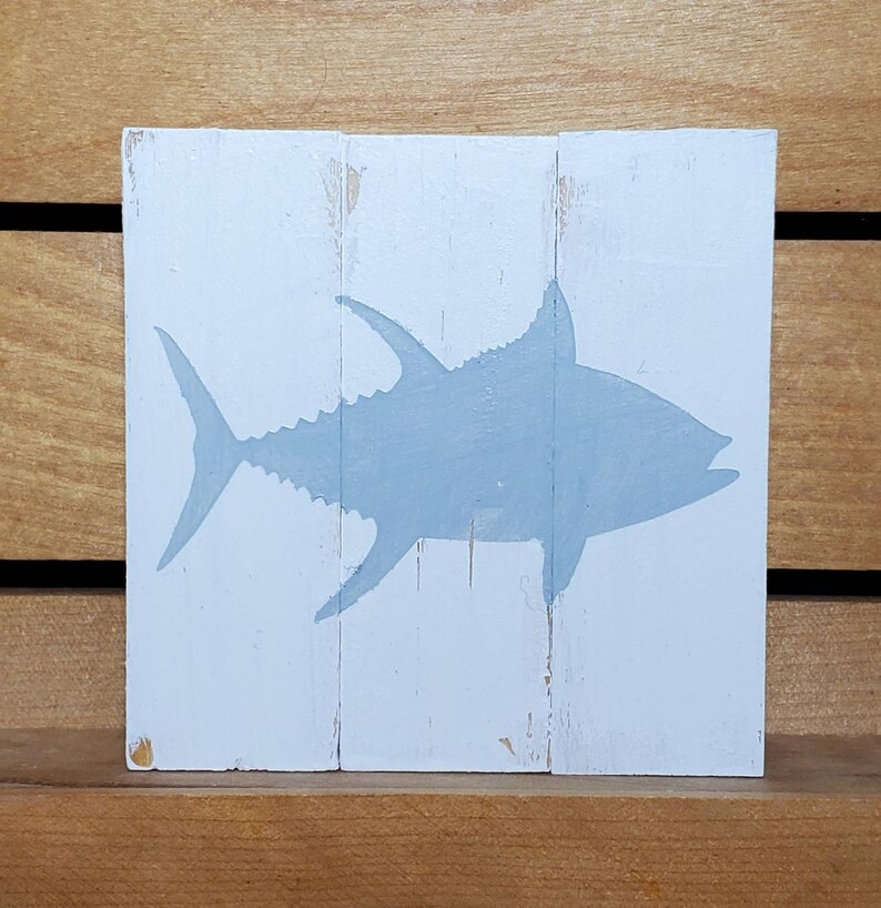 May include: A light blue silhouette of a tuna fish painted on a white wooden sign. The sign is made of three pieces of wood.
