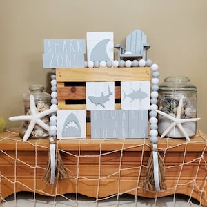 May include: A set of grey wooden signs with shark and ocean-themed designs. The signs read "Shark Zone", "Beware of Sharks", and feature images of sharks. The signs are arranged on a wooden crate with white beads and a small grey chair.
