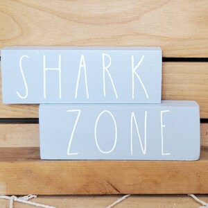 May include: Two light blue wooden blocks stacked on top of each other. The top block says "SHARK" in white paint. The bottom block says "ZONE" in white paint.
