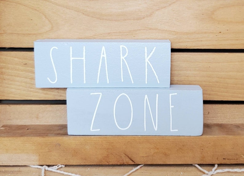 May include: Two light blue wooden blocks stacked on top of each other. The top block says "SHARK" in white paint. The bottom block says "ZONE" in white paint.