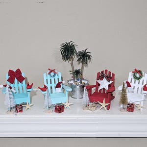 May include: Miniature Adirondack chairs in blue, red, and white, decorated for Christmas. Each chair has festive accents like bows, wreaths, and Santa hats. Small Christmas trees, starfish, and wrapped gifts complete the holiday scene.