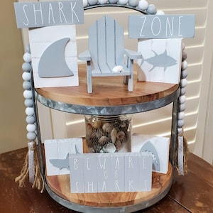 May include: A two-tiered wooden tray with a grey metal rim, decorated with a grey and white beaded garland. The top tier features a miniature grey wooden chair, a grey shark fin block, and a grey fish block with the words "SHARK ZONE". The bottom tier features a glass jar filled with seashells, a grey shark block with the words "BEWARE OF SHARKS", and a grey shark silhouette block.