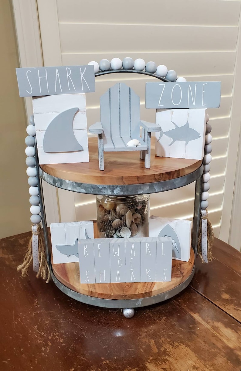 May include: A two-tiered wooden tray with a grey metal rim, decorated with a grey and white beaded garland. The top tier features a miniature grey wooden chair, a grey shark fin block, and a grey fish block with the words "SHARK ZONE". The bottom tier features a glass jar filled with seashells, a grey shark block with the words "BEWARE OF SHARKS", and a grey shark silhouette block.
