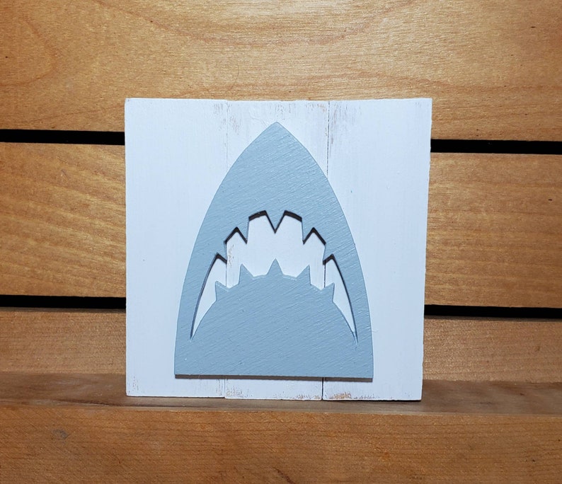 May include: A white wooden sign with a grey shark tooth cutout. The sign is distressed and has a rustic look.