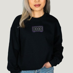 Puede incluir: Sudadera negra con un contorno morado de un reloj digital que muestra "00:00" en la parte delantera. La modelo mide 1,55 m (5'1") y lleva la talla M.