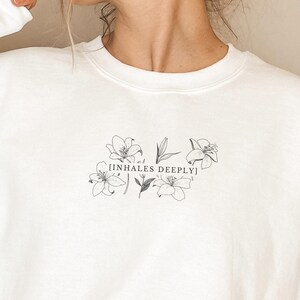 May include: White sweatshirt with a floral design and the text "INHALES DEEPLY" printed on the front.