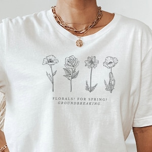Pode incluir: Camiseta branca oversized com um gráfico de quatro flores estilizadas e o texto "FLORALS! FOR SPRING! GROUNDBREAKING."