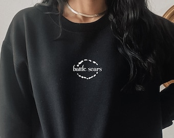 Puede incluir: Sudadera negra de manga larga con un gráfico circular blanco en la parte delantera que dice "battle scars".