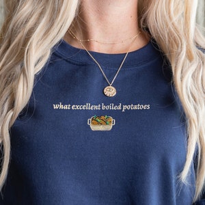 Puede incluir: Una sudadera azul marino con el texto "what excellent boiled potatoes" y una pequeña imagen bordada de un plato de patatas.
