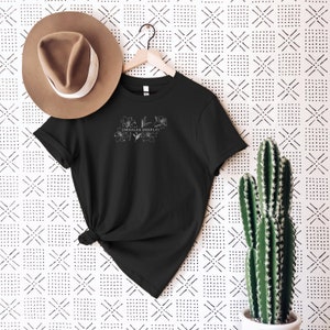 May include: Black t-shirt with a white floral design and the text "inhales deeply" on the front. The shirt is hanging on a hanger against a white background with a black and white geometric pattern.