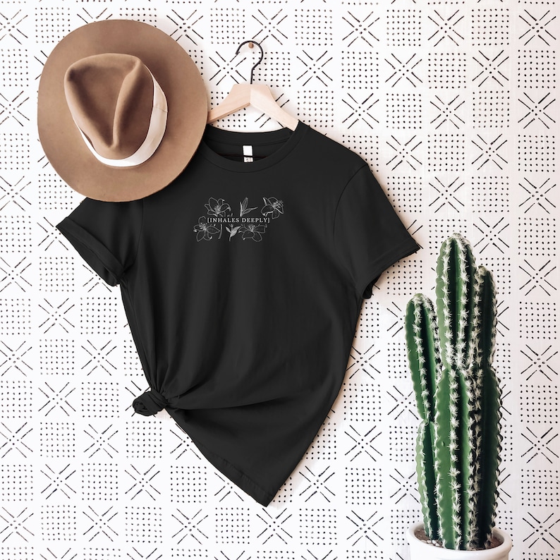 May include: Black t-shirt with a white floral design and the text "inhales deeply" on the front. The shirt is hanging on a hanger against a white background with a black and white geometric pattern.