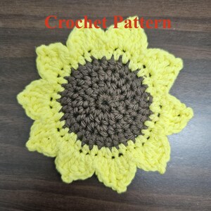 May include: A crocheted sunflower with a brown center and yellow petals. The sunflower is on a brown wooden surface.