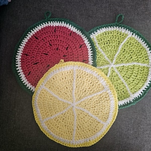 May include: Three crocheted pot holders in the shape of fruit slices: a watermelon, a lime, and a lemon. The watermelon has a red center, white rind, and green edge. The lime is green with white lines, and the lemon is yellow with white lines.
