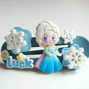 Könnte beinhalten: Ein blauer Plastik-Haarclip mit einem weißen Schneeflockenmuster, einem Cartoon-Bild von Elsa aus Frozen und dem Wort "Luck" in blauen Buchstaben.