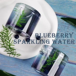 May include: Two clear, cylindrical containers filled with blue liquid and green sprigs, with the text "BLUEBERRY SPARKLING WATER". The containers sit on a white plate and a blue wooden surface, with blueberries scattered around.