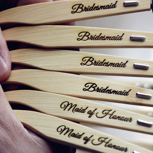 May include: A set of six wooden sunglasses with black lenses. The sunglasses have the words "Bridemaid" and "Maid of Honor" engraved on the wooden frames.