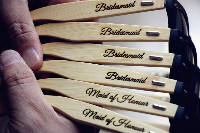 May include: A set of six wooden sunglasses with black lenses. The sunglasses have the words "Bridemaid" and "Maid of Honor" engraved on the wooden frames.