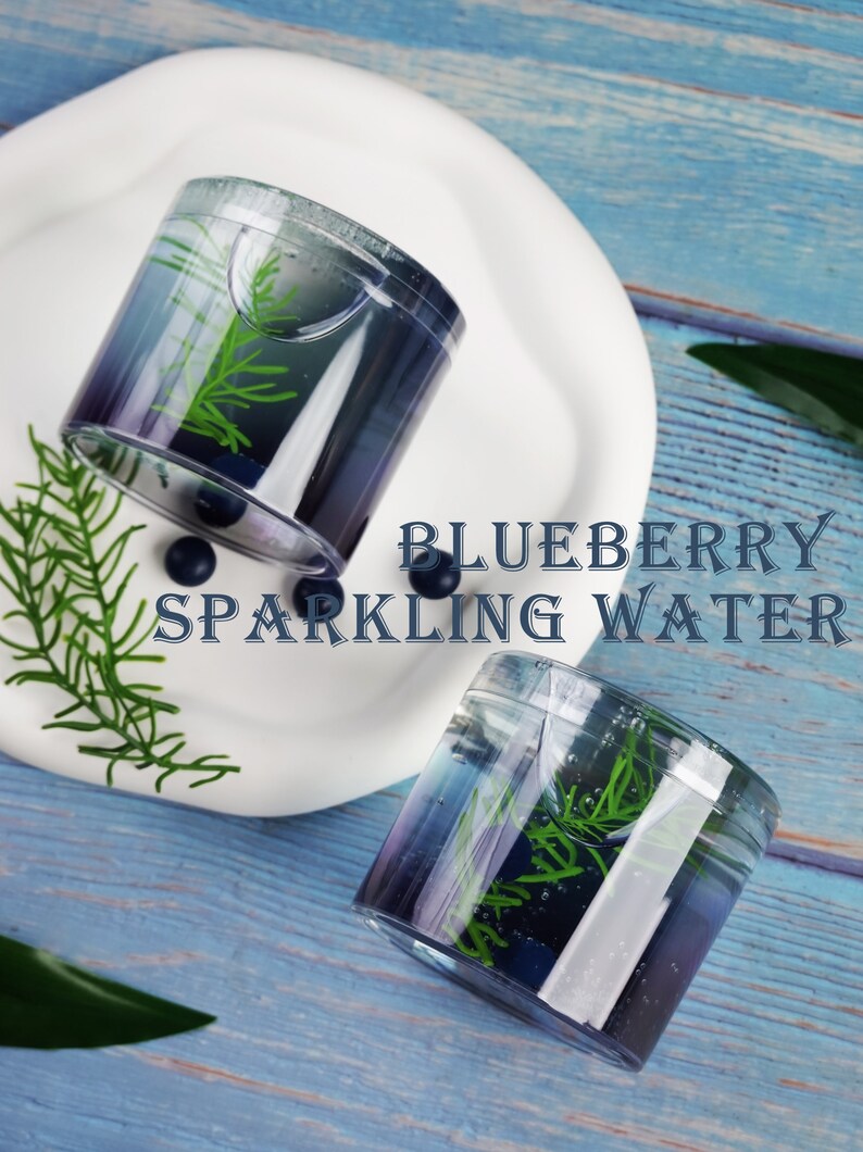 May include: Two clear plastic cups with a gradient blue and purple liquid, containing green sprigs, sit on a white plate. The cups are labeled "BLUEBERRY SPARKLING WATER" in a decorative font. Blueberries and greenery are scattered around the cups on a blue wooden surface.