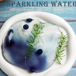 May include: A white plate holds a blue and white liquid with blueberries and green sprigs. The text "BLUEBERRY SPARKLING WATER" is at the top. A basket of blueberries is in the background on a blue wooden surface.
