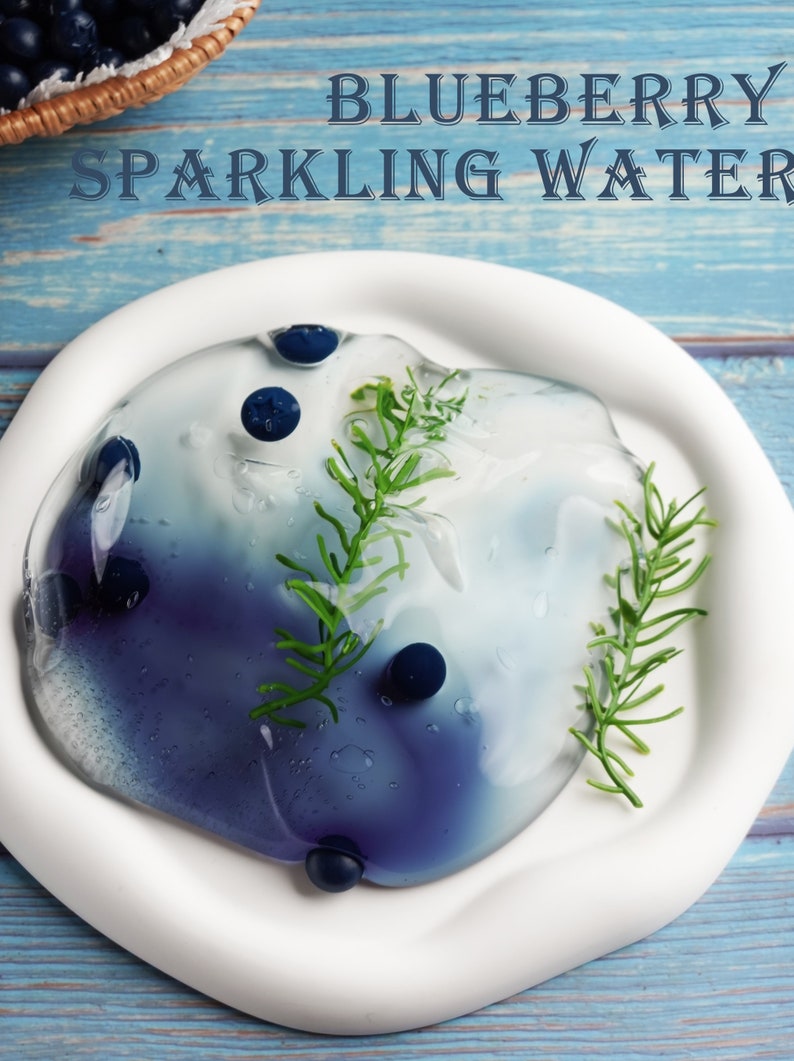 May include: A white plate holds a blue and white liquid with blueberries and green sprigs. The text "BLUEBERRY SPARKLING WATER" is at the top. A basket of blueberries is in the background on a blue wooden surface.
