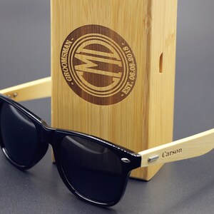May include: Black sunglasses with a bamboo frame and a wooden box. The box is engraved with the text "Groomsman" and "CM" in a circle. The sunglasses have the name "Carson" engraved on the frame.
