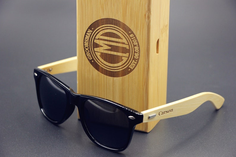 May include: Black sunglasses with a bamboo frame and a wooden box. The box is engraved with the text "Groomsman" and "CM" in a circle. The sunglasses have the name "Carson" engraved on the frame.