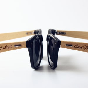 May include: Two pairs of black sunglasses with bamboo frames. One frame is engraved with "Godfather" and the other with "Ariel Marie's".