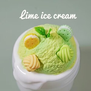 May include: A scoop of lime ice cream in a white bowl, decorated with small, colorful, edible-looking candies. The ice cream is a vibrant green color, and the candies are yellow, green, and cream-colored. The text "lime ice cream" is written in white script.