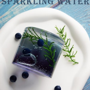 May include: A refreshing glass of blueberry sparkling water, garnished with fresh blueberries and greenery, sits on a white plate. The image features the text "BLUEBERRY SPARKLING WATER" in a stylized font, set against a blue wooden background.