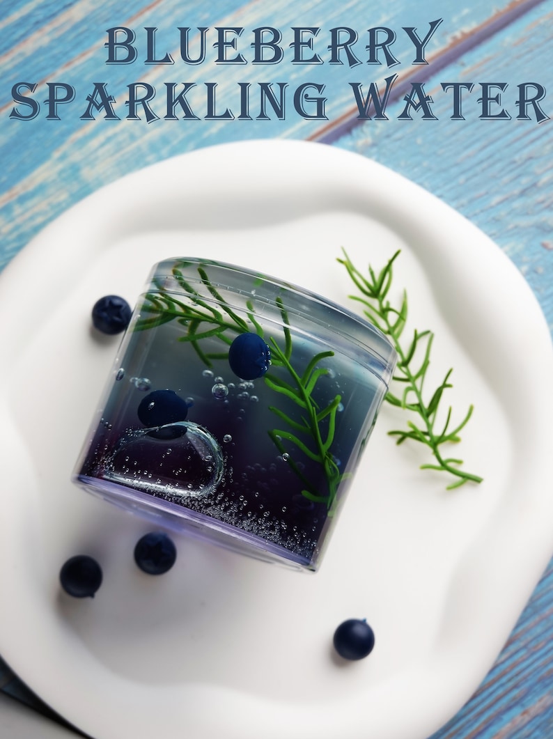 May include: A refreshing glass of blueberry sparkling water, garnished with fresh blueberries and greenery, sits on a white plate. The image features the text "BLUEBERRY SPARKLING WATER" in a stylized font, set against a blue wooden background.