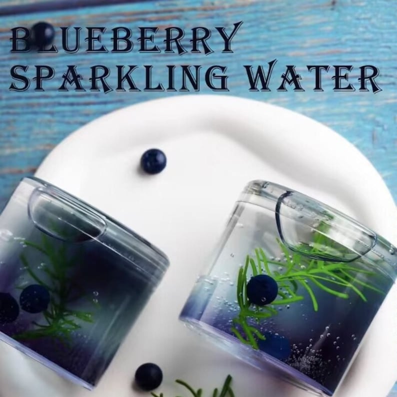 May include: Two clear square glasses filled with blue liquid, blueberries, and green sprigs, sit on a white plate. The text "BLUEBERRY SPARKLING WATER" is above the glasses. The background is a light blue wooden surface.
