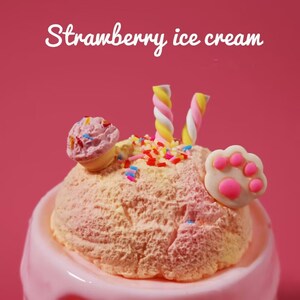 May include: A scoop of strawberry ice cream in a pink bowl, topped with a mini ice cream cone, colorful sprinkles, and two twisted marshmallows. A white paw-shaped decoration with pink pads is also on the ice cream. The words "Strawberry ice cream" are written in white.
